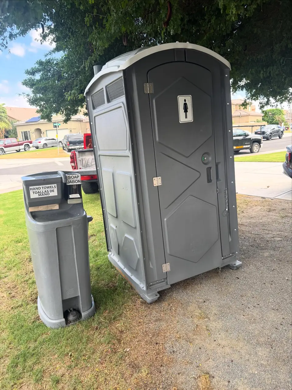 Gray deluxe portable toilet next to a freestanding handwashing sink on a lawn