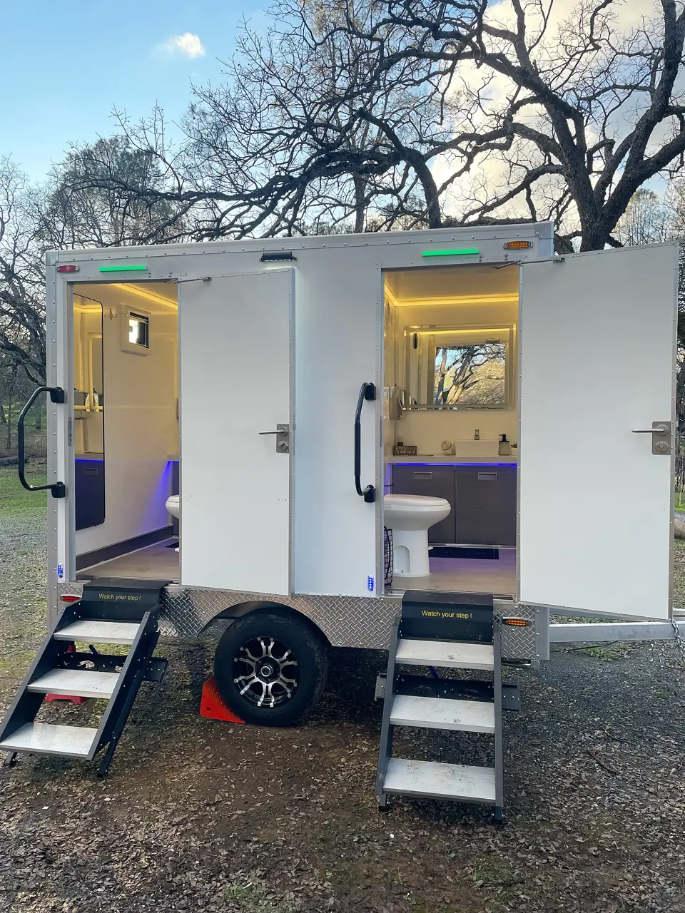 Two-stall restroom trailer with lit interiors and fold-down steps at dusk