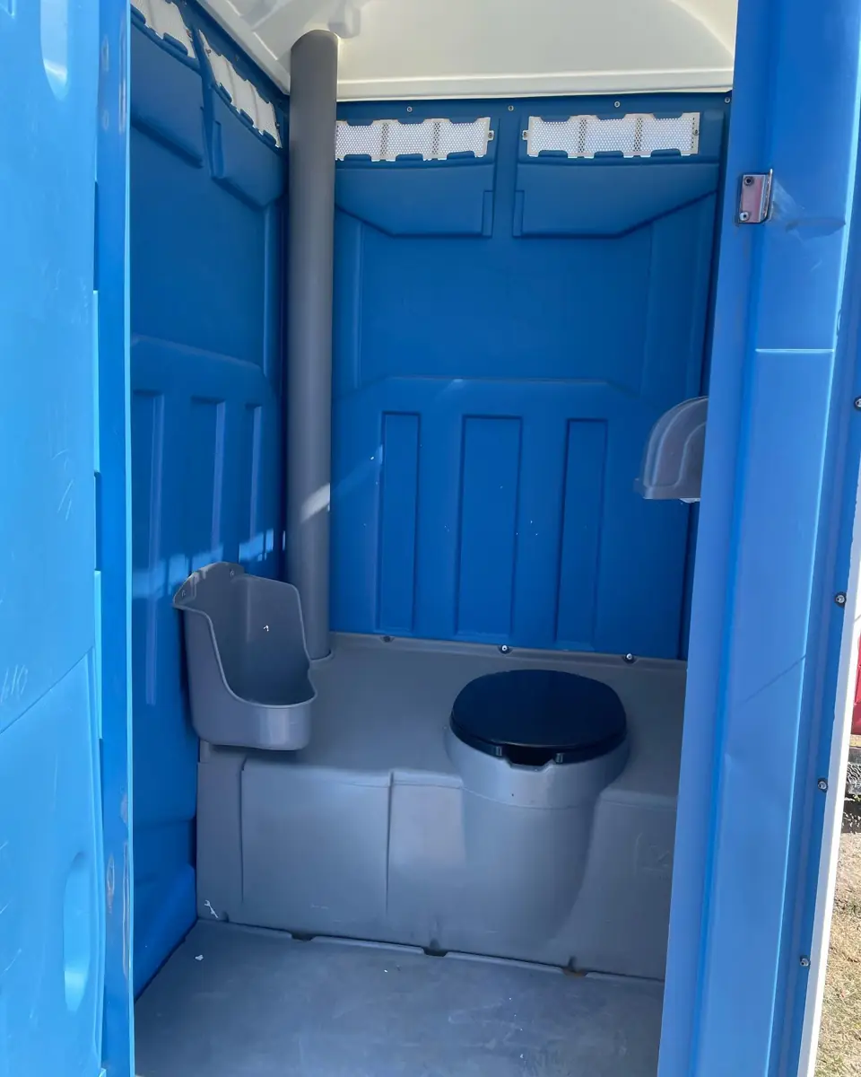 Inside a blue standard porta potty showing the urinal, toilet seat and vent pipe